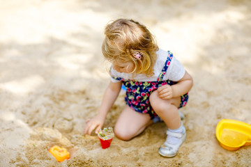 Cute toddler girl playing in sand on outdoor playground. Beautiful baby in red trousers having fun...