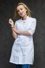 Portrait of a knee-high pretty interesting beautiful girl blonde doctor on a gray background in a white medical coat. Standing in front of the camera, smiling, a lot of emotions.