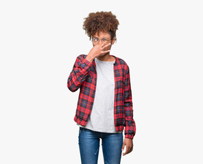 Beautiful young african american woman wearing glasses over isolated background smelling something...