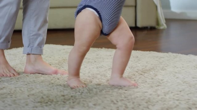 Tilting close-up shot of blond Caucasian toddler just learning to walk on shaggy beige carpet, with help and support from his mother, who is holding him up by both arms raised above head