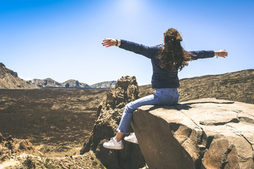 Teenager girl enjoying outdoors sitting on a mountain cliff on the Tenerife Teide National Park. Carefree young girl raised arms  looking at the panorama.  Adventure brave carefree and youth concept.
