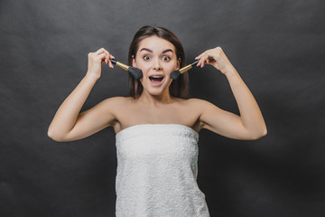 Obraz premium Astonished cute brunette girl in white towel with make up brushes. Isolated on black background.