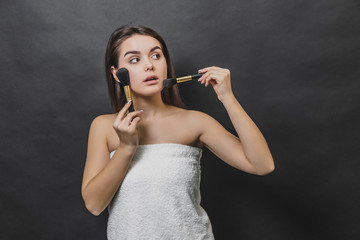 Thoughtful gorgeous girl in white towel holding two makeup brushes. She is looking up and leftwards.