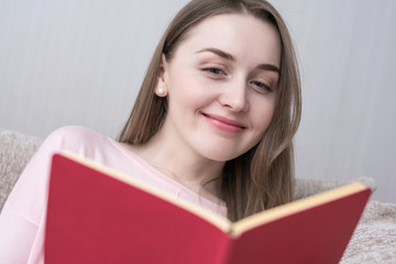 Attractive young girl smiling and reading a book, sitting at home on the couch, close up