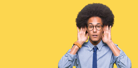 Young african american business man with afro hair wearing glasses Trying to hear both hands on ear gesture, curious for gossip. Hearing problem, deaf