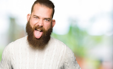 Fototapeta premium Young hipster man wearing winter sweater sticking tongue out happy with funny expression. Emotion concept.