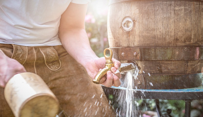 bayerischer Mann in Lederhose sticht ein Holzfass Bier im Garten an und genießt den ersten Schluck