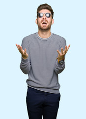 Naklejka premium Young handsome man wearing sunglasses crazy and mad shouting and yelling with aggressive expression and arms raised. Frustration concept.