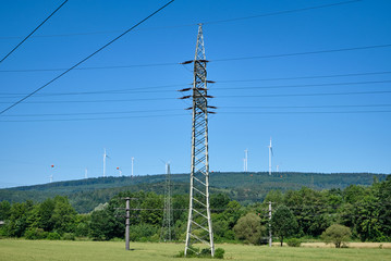 A high electricity pylon against the blue sky in a beautiful rural summer landscape with wind turbines in the background on a hill in the Spessart, Germany