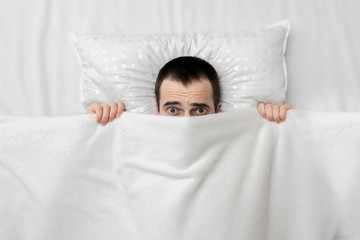 The man woke up in the bedroom, fearfully looking at the camera, hiding under white blanket, top view, copy space