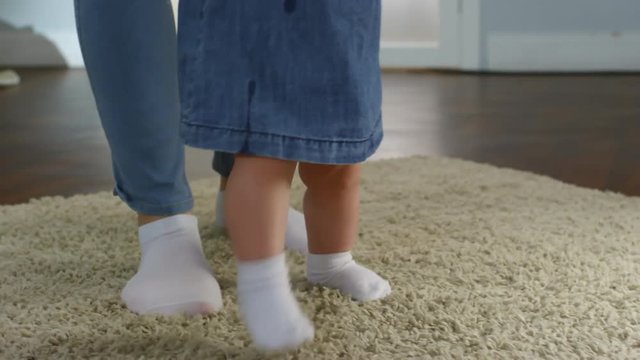 Close-up legs shot of baby girl making unsteady first steps with mother&rsquo;s support on shaggy beige carpet at home, both wearing denim clothes and white socks