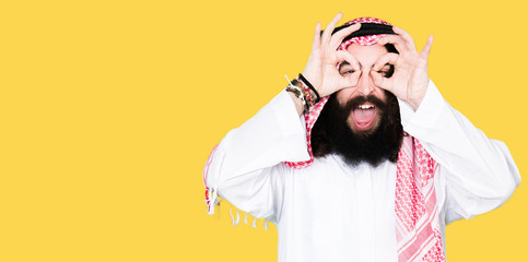 Arabian business man with long hair wearing traditional keffiyeh scarf doing ok gesture like binoculars sticking tongue out, eyes looking through fingers. Crazy expression.