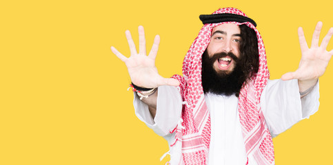 Arabian business man with long hair wearing traditional keffiyeh scarf showing and pointing up with fingers number ten while smiling confident and happy.