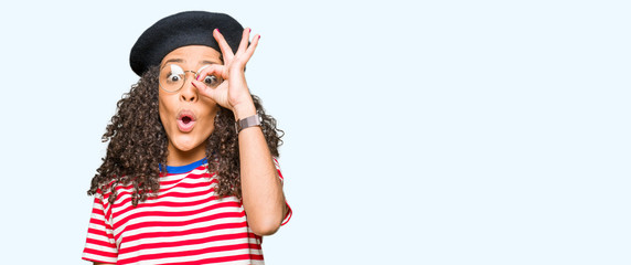 Fototapeta na wymiar Young beautiful woman with curly hair wearing glasses and fashion beret doing ok gesture shocked with surprised face, eye looking through fingers. Unbelieving expression.
