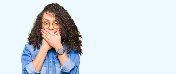Young beautiful woman with curly hair wearing glasses shocked covering mouth with hands for mistake. Secret concept.