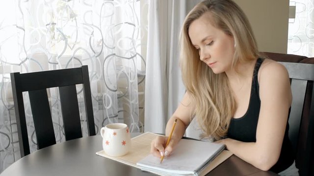Young pregnant blonde is writing in notebook sitting at table in apartment.