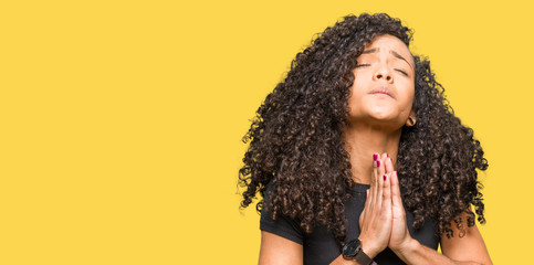 Young beautiful woman with curly hair begging and praying with hands together with hope expression...