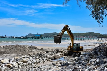 Fototapeta premium Crawler excavator are moving rocks to make breakwater on the beach. The beach , mountains and blue sky blackground