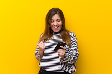 Young girl over yellow wall with phone in victory position