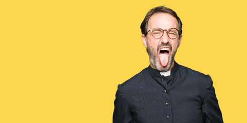 Middle age priest man wearing catholic robe sticking tongue out happy with funny expression. Emotion concept. - obrazy, fototapety, plakaty