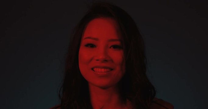 Charming young Asian woman looking to the camera with a smile on the dark background and in the red neon light. Close up. Portrait.