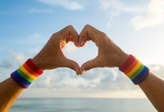 Hands wearing LGBTQI pride rainbow colors sport wristbands making a heart gesture above a golden sunset sea horizon