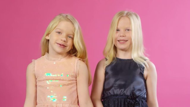 Waist-up shot of lovely 6-year-old blonde Caucasian twin girls with long hair, wearing sparkly party dresses, standing together against pink background in studio, smiling and blowing kisses for camera