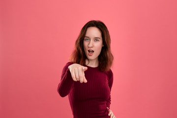 Happy young lady standing isolated over dark pink background looks at the camera while pointing at you expressing emotion of surprise