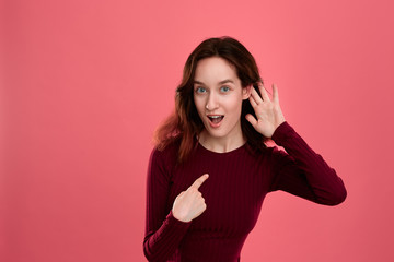 Surprised young lady standing isolated over dark pink background with hands raised close to ear as...