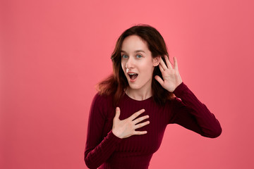 Surprised young lady standing isolated over dark pink background with hands raised close to ear as...