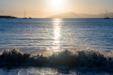 Backlight seascape with a wave crashing on a beach, moored sailboats and the coastline on the...
