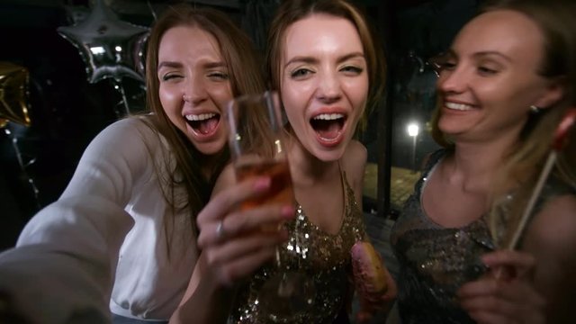 Waist-up handheld selfie shot of three happy slightly tipsy carefree girlfriends dancing at party, gyrating energetically, showing off, taking pictures and posing with glass of champagne and doughnut