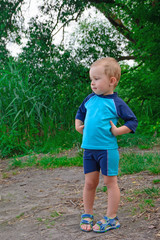 Little boy 3 years old is standing near the water, on the lake shore, dressed in swimming shorts and T-shirt, smiling. Beautiful nature. Space for text