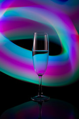 A glass with a drink on the background of a light wave. A single wine glass isolated on a black background with red white pink and orange neon light painting streaks of light behind them.