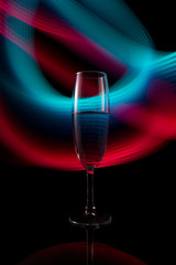 A glass with a drink on the background of a light wave. A single wine glass isolated on a black background with red white pink and orange neon light painting streaks of light behind them.