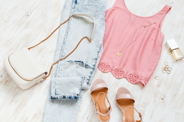 Pink top, blue jeans, small white cross body bag with chain strap, pink heeled sandals on white wooden background. Woman's casual outfit. Trendy spring summer look, pastel colors. Flat lay, top view.