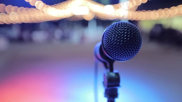 Close-up view on black microphone on stage. Background is out of focus. Thare are many little yellow logts on trop. Pink, blue, violet discolighrts on dancefloor. No people