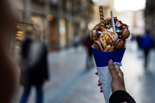 Woman hand holding a bubble waffle with ice cream and candies on a blue paper cone with blurred unrecognizable crowd on the background