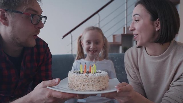 Mother and father holding small birthday cake while daughter blo