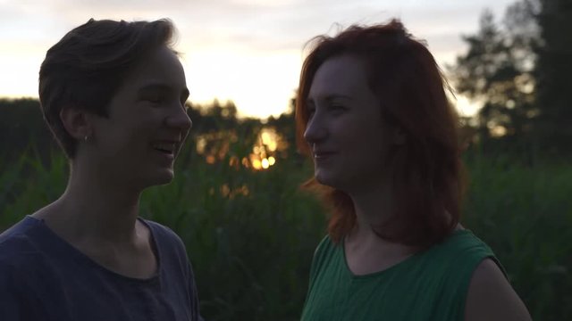 Two women chatting and enjoying sunset in front of high grass - Caucasian white girls wearing a dotted summer dress and a green plain one in indoors