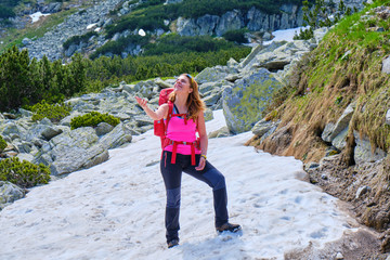 Naklejka premium Happy woman throwing up in the air a snowball, wearing a fuchsia shirt, with a heavy, red backpack, during a Summer hiking tour in Retezat mountains (part of Carpathians), Romania.