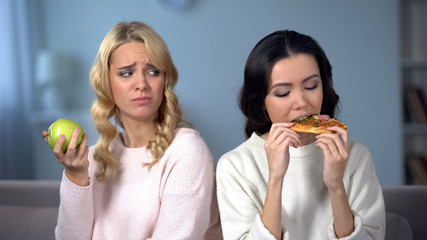 Lady eating pizza while female friend enjoying green apple, healthy diet choice