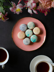 multi-colored macarons on the pink plate on a dark table. white cup of tea or coffee. breakfast with dessert. bouquet of flowers