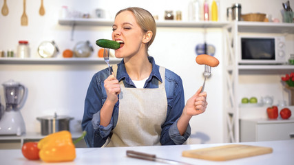 Pretty girl choosing cucumber instead sausage, low-calorie food vs carbohydrates