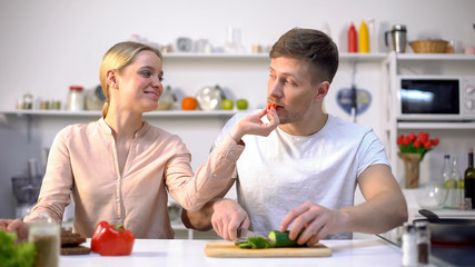 Young happy family flirting, wife feeding husband with raw vegetables, eco food
