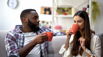 Nice mixed-race couple talking at home, drinking tea after work day, relations