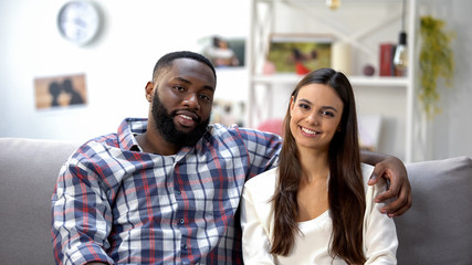 Smiling mixed-race couple sitting on sofa at home and looking at camera, family