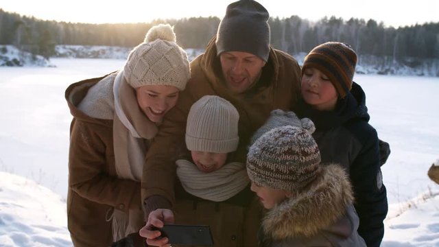 Close-up shot of happy loving family with four kids checking out and admiring their photos on smartphone, scrolling through images and laughing together, with snowy plain and forest in background