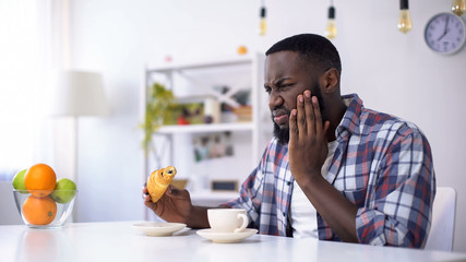 Black man feeling tooth ache from sweet food, eating croissant with coffee - obrazy, fototapety, plakaty