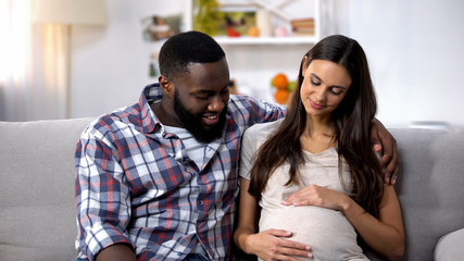 Mixed-race family sitting on sofa, pregnant woman holding tummy, happiness